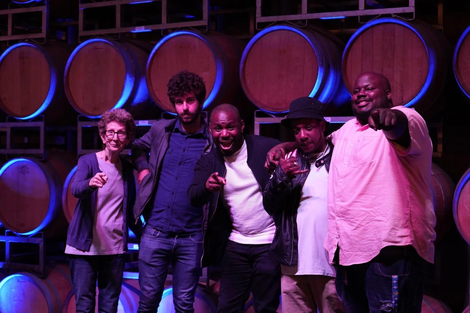 Five comedians posing together in front of wine barrels